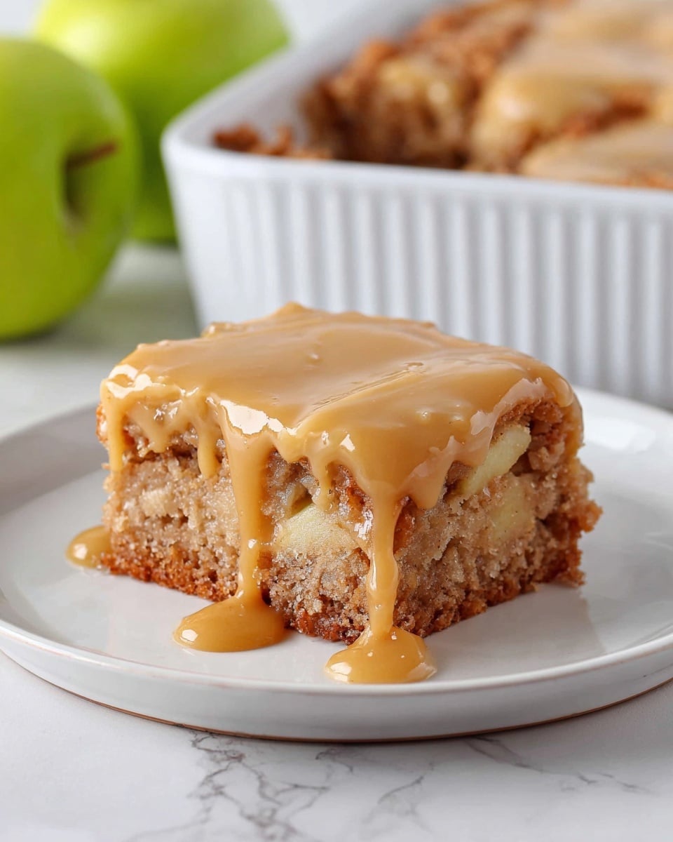 A square piece of dessert sits on a white plate placed on a white marbled surface. The dessert has two visible layers: the bottom layer is a soft, crumbly cake with a golden brown color and rough texture, showing bits of apple inside, while the top layer is a smooth, thick caramel-colored icing that drips slightly down the sides. In the background, a white baking dish with more of the dessert and a green apple can be seen, adding contrast to the warm tones of the dessert. Photo taken with an iphone --ar 4:5 --v 7
