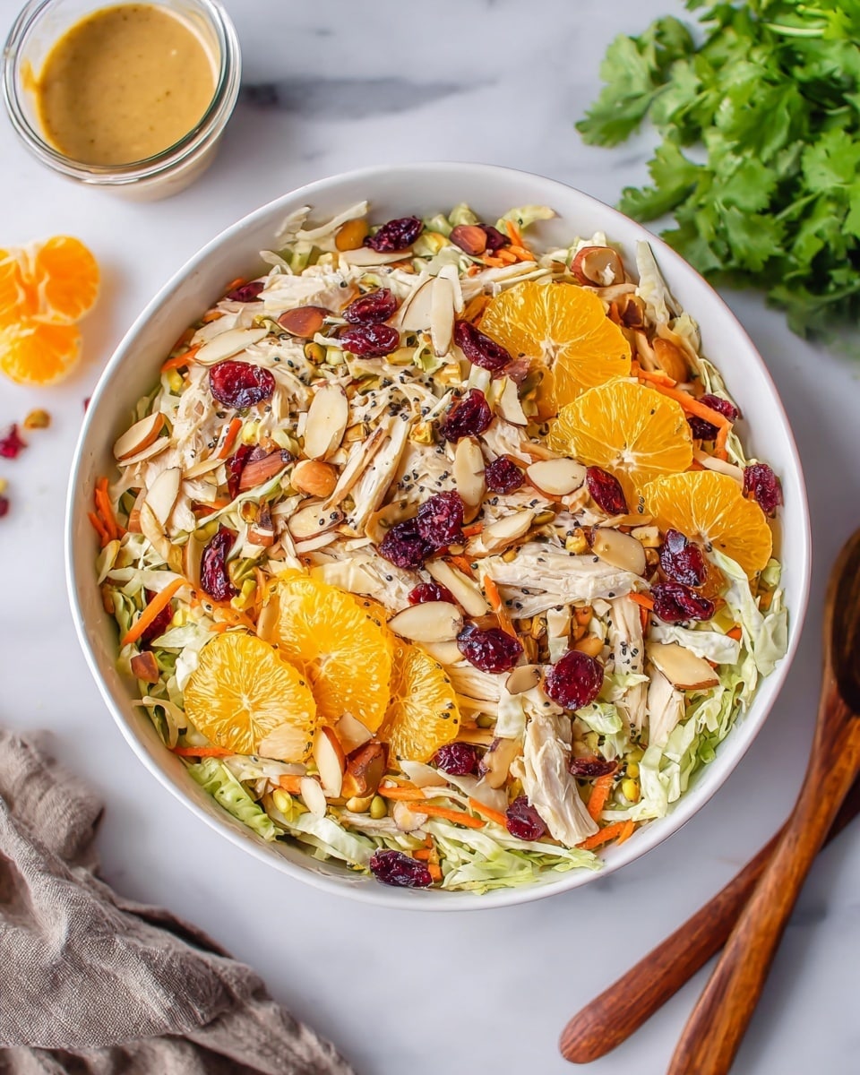 A close-up view of a white bowl filled with a fresh layered salad on a white marbled texture background, showing a mix of shredded light green cabbage and orange carrot strips as the base layer. Scattered on top are bright orange mandarin slices, deep red dried cranberries, and light brown almond slices. Small white chunks of cooked chicken and some black poppy seeds are mixed throughout, adding texture and contrast. To the top left of the bowl, there is a small glass container of light brown dressing. A bunch of fresh green cilantro sits in the top right corner, and wooden salad utensils rest on the bottom right on the same white marbled surface. Photo taken with an iphone --ar 4:5 --v 7