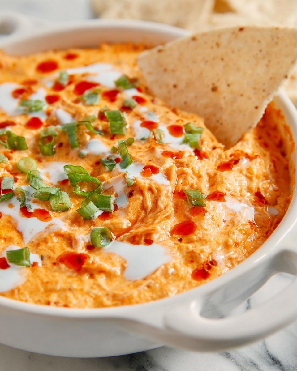 The image shows a close-up of a creamy orange dip in a white bowl with handles. The dip has a smooth texture with bits of shredded ingredients, topped with a drizzle of white sauce and small drops of red sauce. It is also sprinkled with chopped green herbs or scallions, adding a pop of green color on top. A crisp light beige chip is partially dipped into the sauce, resting near the center of the bowl. The background features a white marbled surface. photo taken with an iphone --ar 4:5 --v 7