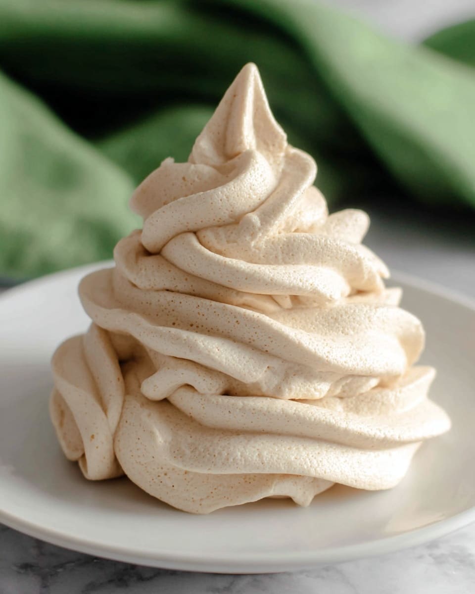 A swirl of creamy beige frosting is piped in four thick, smooth layers, each with soft peaks and gentle curves twisting upwards to form a tall, pointed top. The texture looks light and fluffy, with tiny air holes visible throughout. This frosting mound sits centered on a plain white plate, placed on a white marbled surface with a soft green fabric blurred in the background. photo taken with an iphone --ar 4:5 --v 7