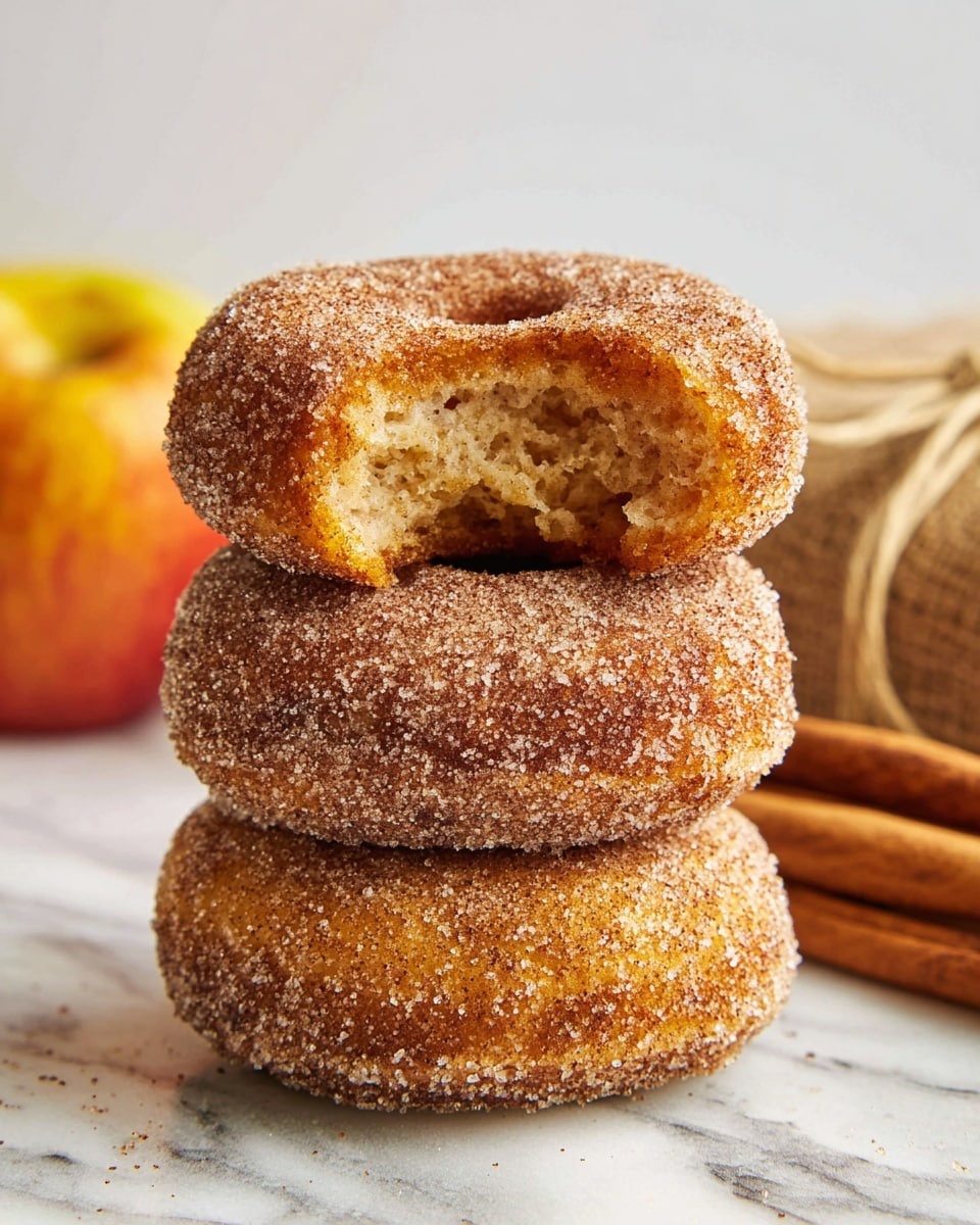 A stack of three sugar-coated doughnuts is shown on a white marbled textured surface, with the top doughnut having a bite taken out to reveal a soft, light brown inside with air pockets. The doughnuts are round and thick, each covered evenly with a layer of granulated sugar mixed with cinnamon, giving them a slightly rough texture and a sparkling appearance. The middle doughnut is a deeper brown color, while the bottom one appears lighter. In the blurred background, there is a bundle of cinnamon sticks tied with twine and a partially visible yellow and red apple. The overall lighting is bright and natural, highlighting the texture and color contrast of the doughnuts. photo taken with an iphone --ar 4:5 --v 7