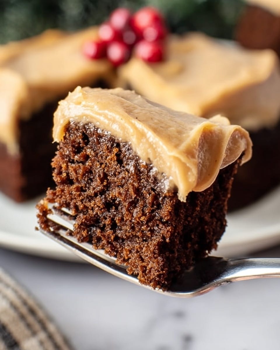A close-up view of a single slice of moist, dense dark brown cake with a thick layer of smooth, light tan frosting evenly spread on top; the frosting has a creamy texture with gentle swirls, and the cake's crumb is visible with small, even holes. The background shows blurred red berries and green leaves, resting on a white marbled surface. photo taken with an iphone --ar 4:5 --v 7