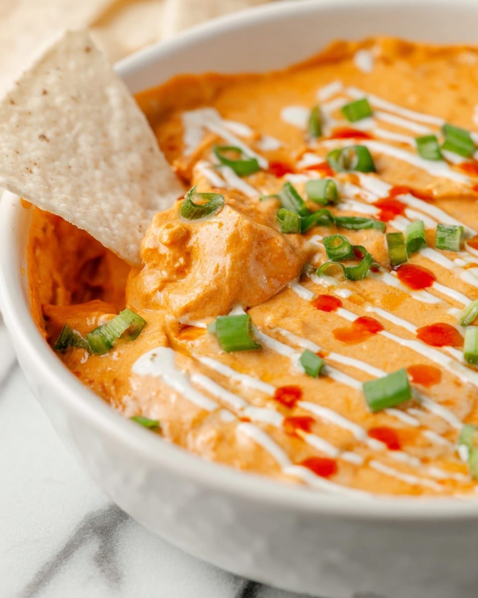 A close-up view of a creamy orange dip in a white bowl with a smooth, slightly chunky texture; the dip is decorated with thin white sauce lines and small red sauce drops on top. There are scattered chopped bright green onions on the surface, adding color contrast. A white tortilla chip is dipped partially into the creamy mixture on the left side of the bowl. The background shows a white marbled texture. photo taken with an iphone --ar 4:5 --v 7