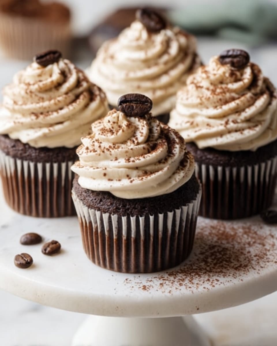 The image shows four chocolate cupcakes on a white cake stand with a white marbled surface below. Each cupcake has a dark brown base and is topped with a swirl of light brown cream with a dusting of cocoa powder. On top of each cream swirl, there is a single dark coffee bean. Scattered coffee beans are placed around the cupcakes on the cake stand. The background is softly blurred, keeping the focus on the cupcakes. photo taken with an iphone --ar 4:5 --v 7