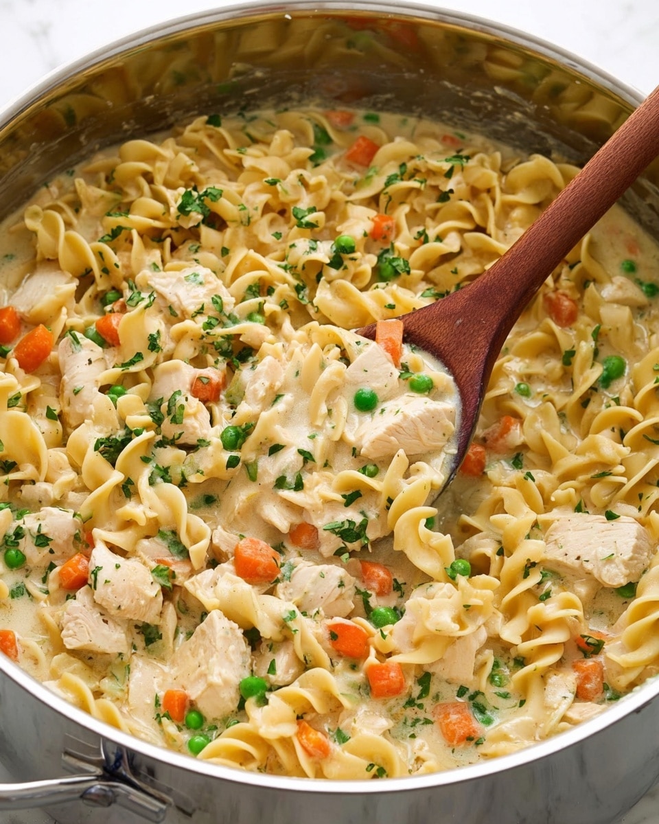 A close-up view of a large silver pot filled with creamy chicken noodle soup. The dish has three layers: the base layer is a pale creamy sauce, topped with thick, curly yellow egg noodles scattered all over. Pieces of cooked white chicken chunks, small cubes of orange carrots, and bright green peas are mixed throughout, along with fresh chopped green herbs sprinkled on top. A wooden spoon rests inside the pot, lightly lifting some of the soup, showing the thick texture of the sauce and bits of ingredients. The background is a white marbled surface. photo taken with an iphone --ar 4:5 --v 7