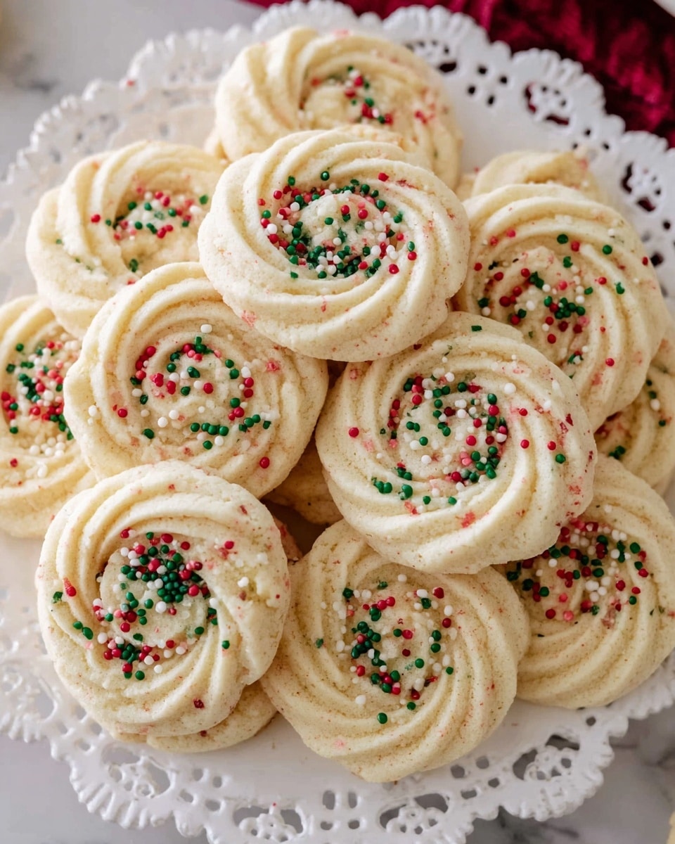 A pile of light cream-colored swirl cookies with a soft, crumbly texture sits on a white plate with a lace edge. Each cookie has one layer, shaped in a round spiral pattern with ridges, and several are sprinkled with small round sprinkles in red, green, and white colors spread unevenly on top. The plate rests on a white marbled textured surface. photo taken with an iphone --ar 4:5 --v 7