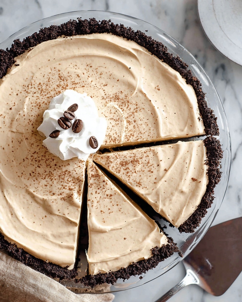 The image shows a creamy coffee pie in a glass pie dish with a dark chocolate crumb crust forming the first layer around the edges. The second layer is a smooth, light beige coffee-flavored cream spread thick and evenly across the crust with gentle swirls on the surface. A dollop of white whipped cream sits on one side of the pie's top, garnished with three dark coffee beans and sprinkled lightly with coffee powder over the entire pie. Three slices have been cut, showing the separation between pieces. The pie dish sits on a white marbled surface with a beige cloth in the corner and a silver pie server nearby. Photo taken with an iphone --ar 4:5 --v 7