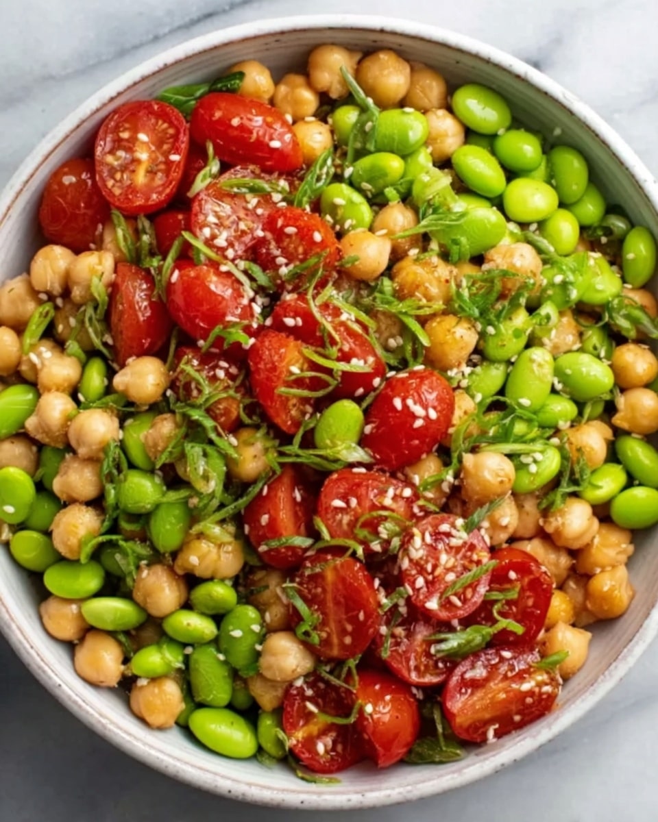 A white bowl filled with a colorful chickpea salad sits on a white marbled surface. The salad has several layers including bright green edamame beans scattered throughout, soft beige chickpeas, and shiny, halved red cherry tomatoes. Thinly sliced green onions are sprinkled all over, adding a fresh touch. The textures vary from smooth chickpeas to firm edamame and juicy tomatoes, with some sesame seeds visible on top, enhancing the visual appeal. photo taken with an iphone --ar 4:5 --v 7