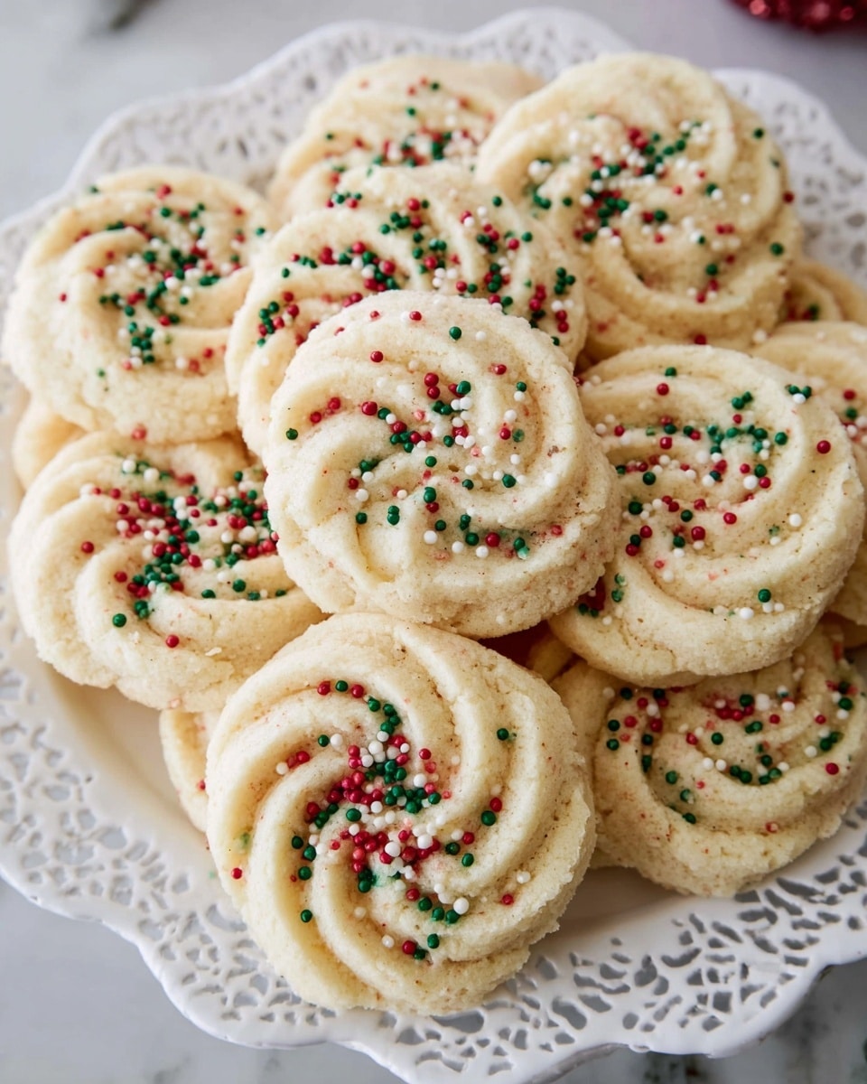 A white plate filled with about a dozen soft, pale yellow cookies shaped like roses with thick, swirling layers, some topped with small round sprinkles in red, green, and white colors that stand out against the cookies' smooth, slightly crumbly texture, all set on a white marbled surface. photo taken with an iphone --ar 4:5 --v 7