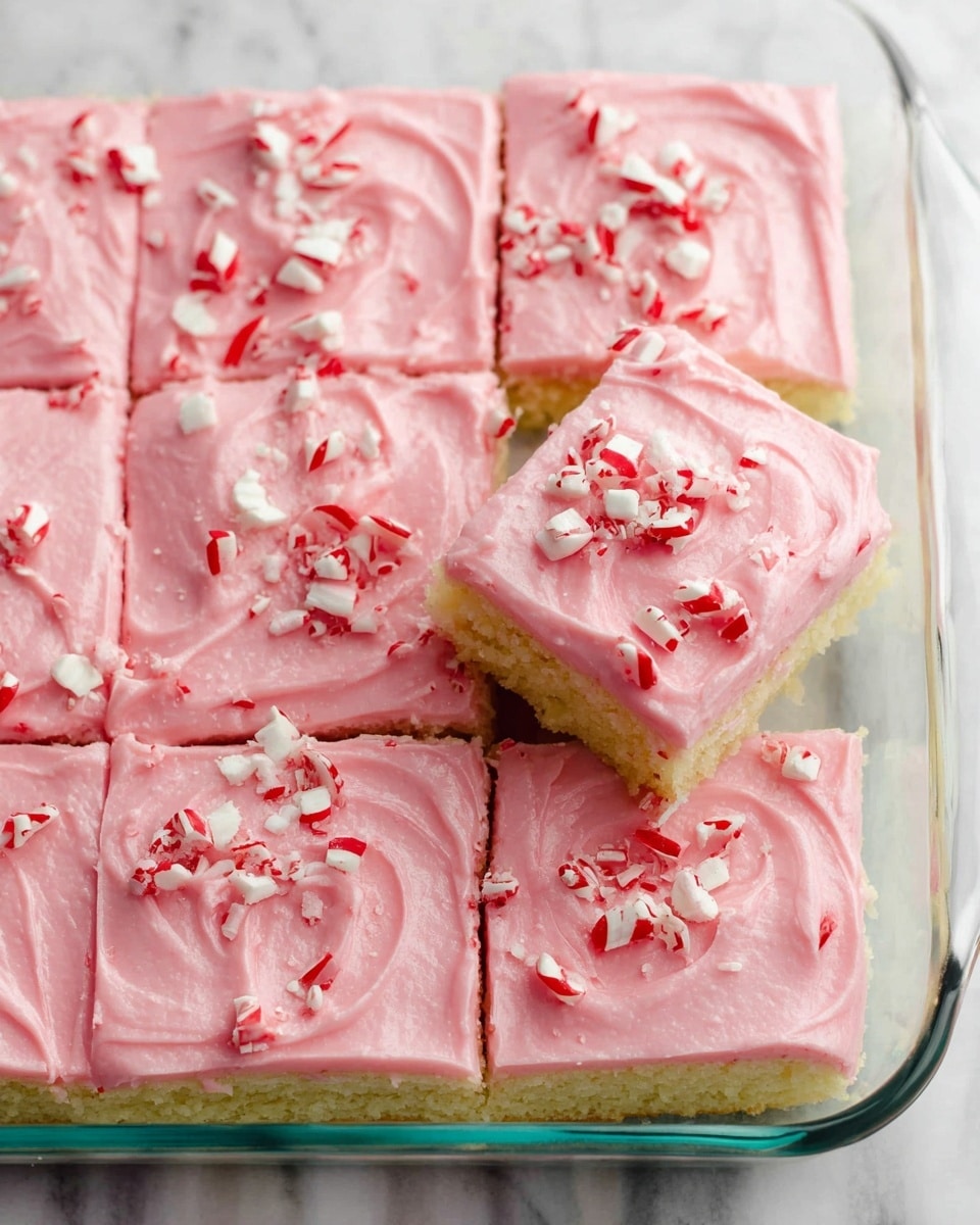 The image shows a dessert cut into nine squares in a clear glass baking dish on a white marbled surface. The dessert has two visible layers: the bottom layer is a pale yellow soft cake, and the top layer is thick, smooth, and creamy pink frosting with a slightly swirled texture. Small pieces of crushed red and white peppermint candy are scattered evenly on top of the pink frosting, adding a festive touch. photo taken with an iphone --ar 4:5 --v 7