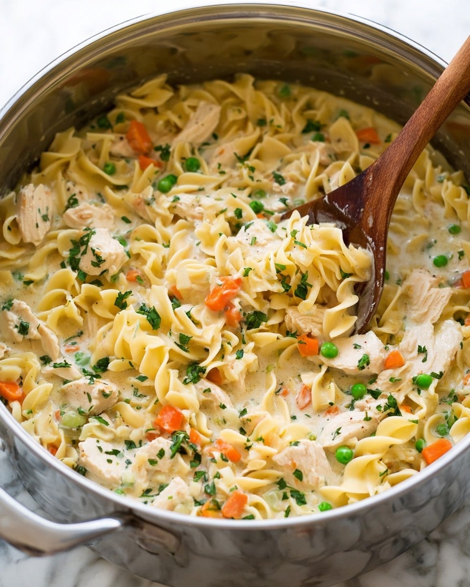 A close-up view of a large silver pot filled with creamy chicken noodle soup. The dish has many layers: the base is pale yellow egg noodles with a curly texture, mixed with small cubes of light orange carrots and bright green peas scattered throughout. Chunks of white, cooked chicken are evenly spread in the pot, all coated in a thick, light beige creamy broth. Small sprigs of fresh green parsley are sprinkled on top, adding a touch of freshness and color contrast. A well-used wooden spoon is partly submerged in the soup, stirring the ingredients gently. The background shows a smooth white marbled texture. Photo taken with an iphone --ar 4:5 --v 7