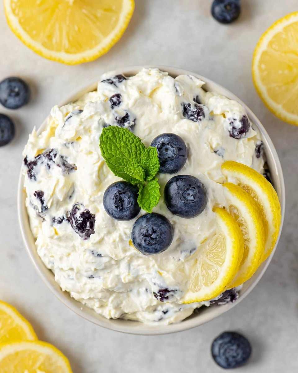 A white bowl filled with a creamy, white mixture dotted with dark blueberries throughout. On top, there are four whole blueberries placed in the center along with two fresh green mint leaves. Two thin lemon slices are placed on the side of the bowl, leaning against the creamy mixture. The bowl rests on a white marbled surface with parts of other bowls and lemon slices visible around it. photo taken with an iphone --ar 4:5 --v 7
