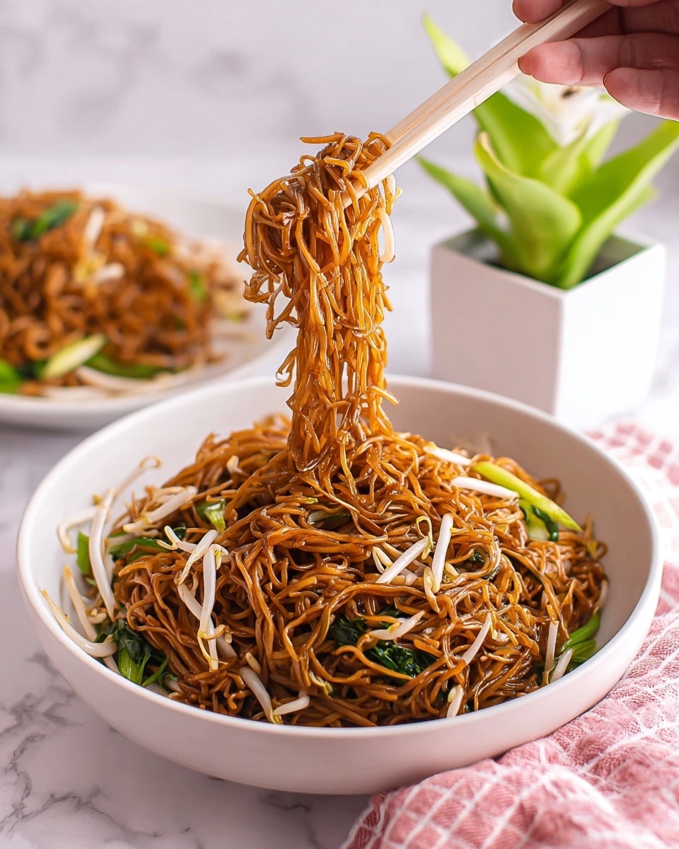 A white bowl filled with a generous serving of dark golden brown fried noodles mixed with green leafy vegetables and pale bean sprouts, with thin, slightly shiny strands piled high in the bowl. A woman's hand is lifting a clump of the noodles with light pink chopsticks, the noodles hanging with some green and white vegetables caught in the strands. In the background, there is another white bowl with more noodles placed on a white marbled surface, and a pink checkered cloth is partly visible at the bottom of the image, along with a green potted plant in a white square pot. Photo taken with an iphone --ar 4:5 --v 7