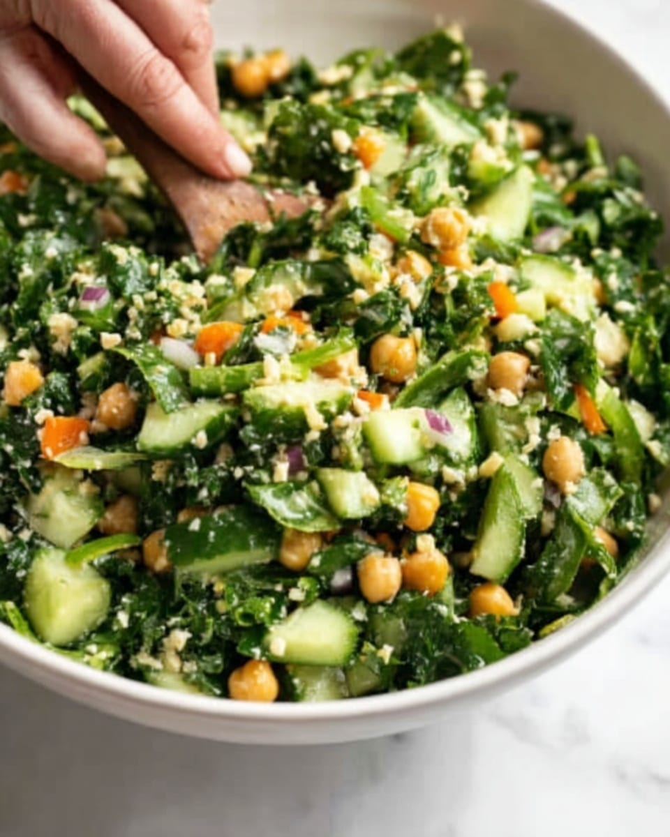 The image shows a fresh green salad in a white bowl. The salad has many layers including small green leafy vegetables, chopped cucumbers, chickpeas, and tiny pieces of orange carrot. The textures look soft and crunchy mixed together, with some grain-like bits spread through the salad. A woman's hand is seen on the left side, gently holding or mixing the salad. The background is a white marbled texture. Photo taken with an iphone --ar 4:5 --v 7
