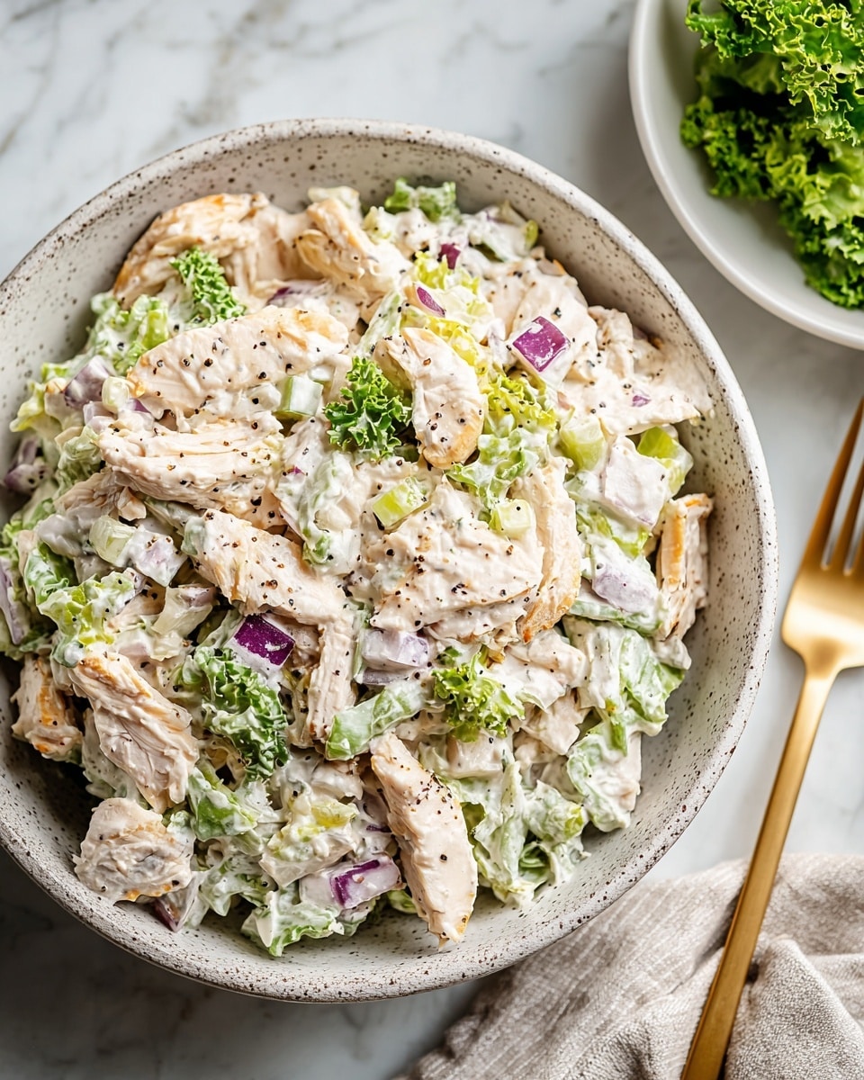 A white speckled bowl holds a chicken salad with visible layers: the base layer is chopped green leafy lettuce, topped with shredded white chicken pieces mixed with creamy dressing that coats the chicken and lettuce evenly; scattered purple onion chunks add color contrast, while small black pepper specks are sprinkled on top. The bowl sits on a white marbled surface, with a golden fork and a bowl of green lettuce blurred in the background. photo taken with an iphone --ar 4:5 --v 7