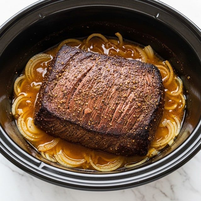 A large piece of cooked roast beef sits in the center of a black slow cooker, resting on a layer of sliced onions soaked in brown cooking juices. The beef has a dark brown, slightly crispy and seasoned outer crust with visible black pepper and herbs, showing some texture and marbling on the surface. The onions beneath are soft and translucent, immersed in the glossy juices that pool around the meat inside the slow cooker. The slow cooker is placed on a white marbled surface. photo taken with an iphone --ar 4:5 --v 7