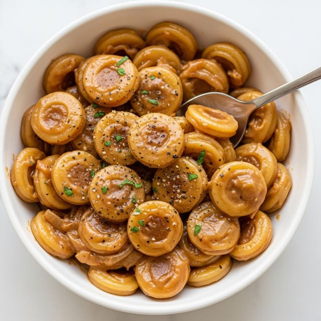 A white bowl is filled with a creamy, brown sauce lightly coating round pasta pieces. The pasta has a glossy texture and is sprinkled with small green herb bits and freshly ground black pepper, giving a fresh and spicy look. A silver spoon is partially digging into the pasta, showing the thick sauce covering the curved edges and flat surfaces of the pasta evenly. The bowl sits on a white marbled surface with a soft background. photo taken with an iphone --ar 4:5 --v 7