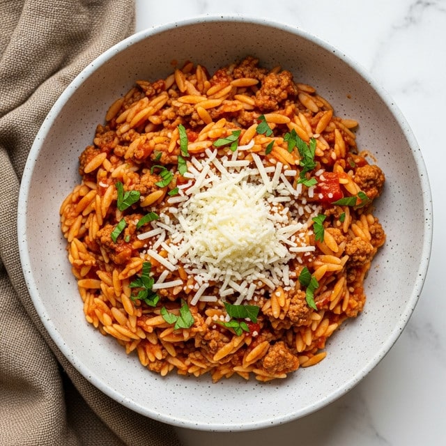 A bowl of orzo pasta cooked in a rich red tomato sauce mixed with browned ground meat, filling the bowl evenly. On top of the orzo is a generous layer of finely grated white cheese scattered mostly in the center, sprinkled with small pieces of fresh green parsley. The bowl is light grey with speckles and sits on a white marbled surface next to a piece of rough brown fabric. Photo taken with an iphone --ar 4:5 --v 7