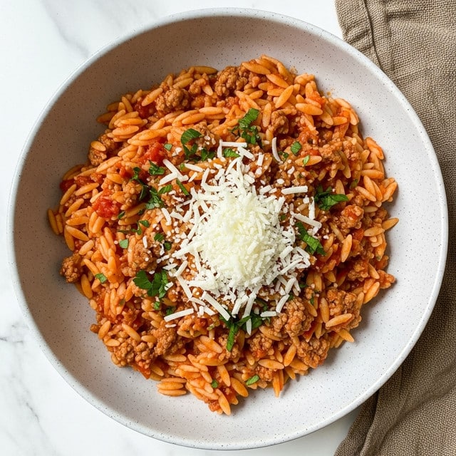 A bowl filled with three main layers starts with a base of small, shiny, light yellow pasta pieces shaped like rice grains, mixed with chunky ground meat in a reddish tomato sauce. On top of this, a layer of finely grated white cheese spreads across the center, with bright green parsley leaves scattered over the cheese and surrounding pasta for color. The bowl is light gray speckled, placed on a rough beige cloth over a white marbled surface. photo taken with an iphone --ar 4:5 --v 7