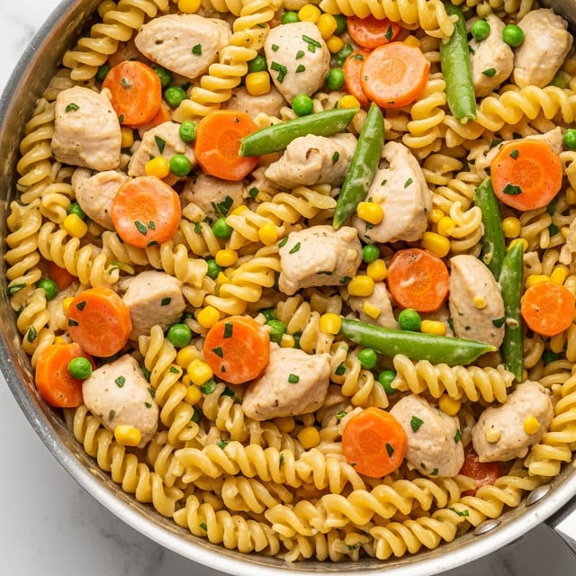 A close-up of a silver pan with creamy pasta, showing three layers of ingredients: at the base, light yellow cooked rotini pasta twisted in spiral shapes; mixed in the middle are small bright green peas, thin round orange carrot slices, and golden yellow corn kernels; scattered on top are chunks of light brown cooked chicken. All the ingredients are coated in a thick creamy sauce with specks of green herbs evenly spread throughout. The pan is placed on a white marbled texture surface. photo taken with an iphone --ar 4:5 --v 7