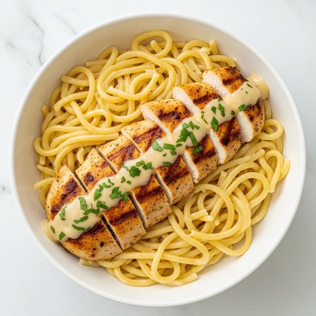 The image shows a white bowl filled with creamy pasta at the bottom, with smooth, light yellow noodles layered evenly. On top, there are several golden brown grilled chicken pieces arranged in a neat row, each piece with a slightly crispy texture and small grill marks. A light creamy sauce is drizzled over the chicken, adding a glossy finish, and sprinkled green herbs are scattered on top, adding a fresh color contrast. The bowl is set on a white marbled surface, making the dish stand out. Photo taken with an iphone --ar 4:5 --v 7