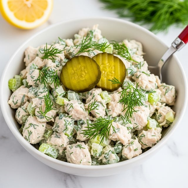 A white bowl filled with creamy chicken salad that has small chunks of white chicken mixed with diced green celery and fresh dill scattered on top, giving a fresh green sprinkle over the pale mix. Two thick green pickle slices rest on top toward one side, adding a bright contrast. A silver spoon with a red handle sits inside the bowl on the right side. The bowl is placed on a white marbled surface with some green leafy herbs and a halved lemon blurred in the background. Photo taken with an iphone --ar 4:5 --v 7