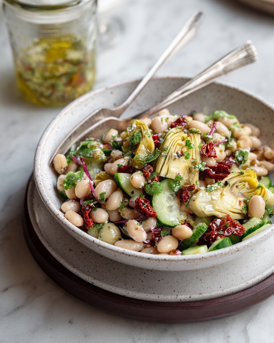 A white speckled bowl filled with a colorful bean salad placed on top of a white plate, which sits on a dark brown plate, all set against a white marbled surface. The salad has three main layers: a base layer of mixed white and red beans with a smooth, slightly shiny texture; scattered over this are slices of bright green cucumber and pale yellow artichoke hearts with soft, fibrous textures; on top, small chopped sun-dried tomatoes add a deep red color and rough texture, along with finely chopped leafy herbs in green that provide a fresh appearance. A silver fork rests inside the bowl, leaning on the edge, and a clear glass jar with dressing sits blurred in the background. Photo taken with an iphone --ar 4:5 --v 7