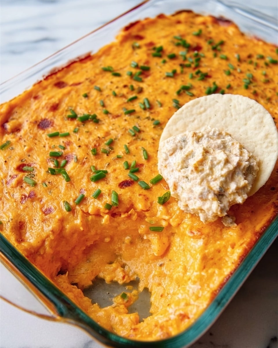 The image shows a glass baking dish filled mostly with a bright orange baked layer that looks creamy and slightly browned on top. The surface is dotted with small green chives, adding a touch of color. On one corner of the dish, a scoop of a chunky, off-white creamy dip with visible small bits is placed, holding a lightly speckled, white round chip standing upright. The dish sits on a white marbled surface. photo taken with an iphone --ar 4:5 --v 7