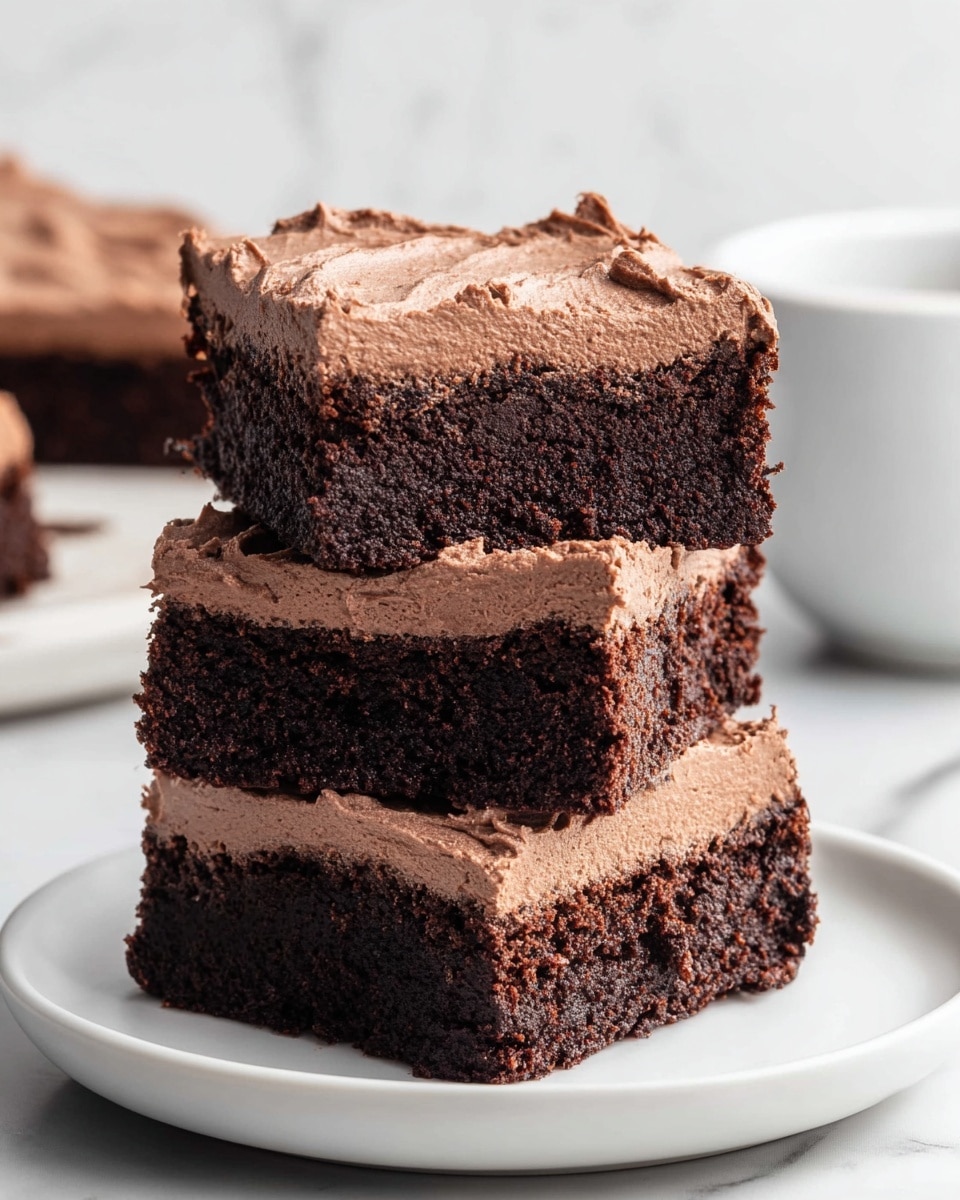 Three square pieces of chocolate brownies are stacked on a white plate, each piece showing two layers: a dense, dark brown brownie base with a slightly crumbly texture, and a thick, creamy light brown chocolate frosting on top with soft swirled patterns. The stack is centered on a white marbled surface with a blurred white cup in the background. Photo taken with an iphone --ar 4:5 --v 7
