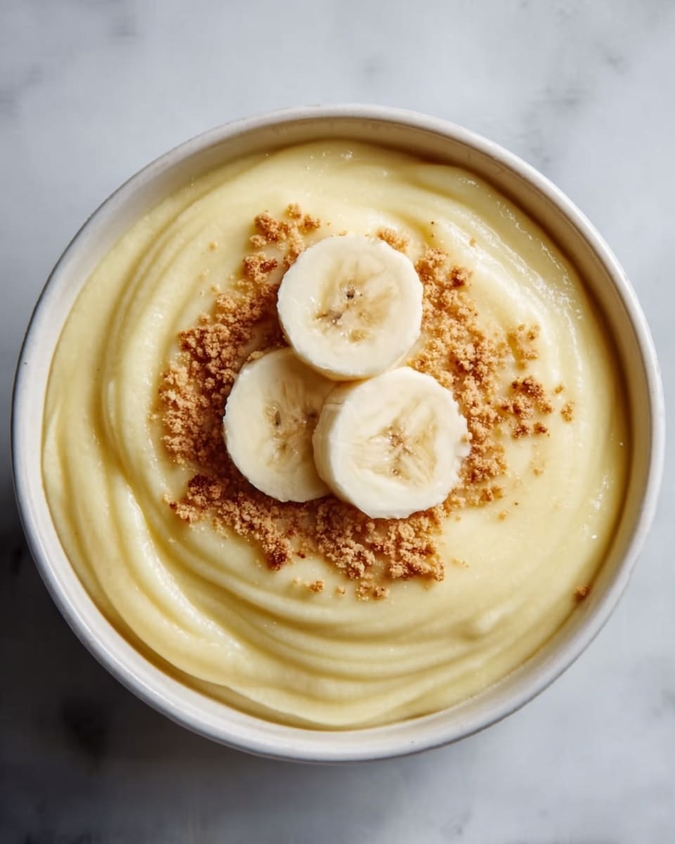 A close-up top view of a white bowl filled with smooth, creamy yellow pudding that is swirled in soft waves. On top, there are three round banana slices arranged in the center, and around them is a sprinkle of light brown crushed nuts or crumbs spread in a circular pattern. The bowl sits on a white marbled textured surface. The lighting is soft and natural, highlighting the pudding’s silky texture. photo taken with an iphone --ar 4:5 --v 7