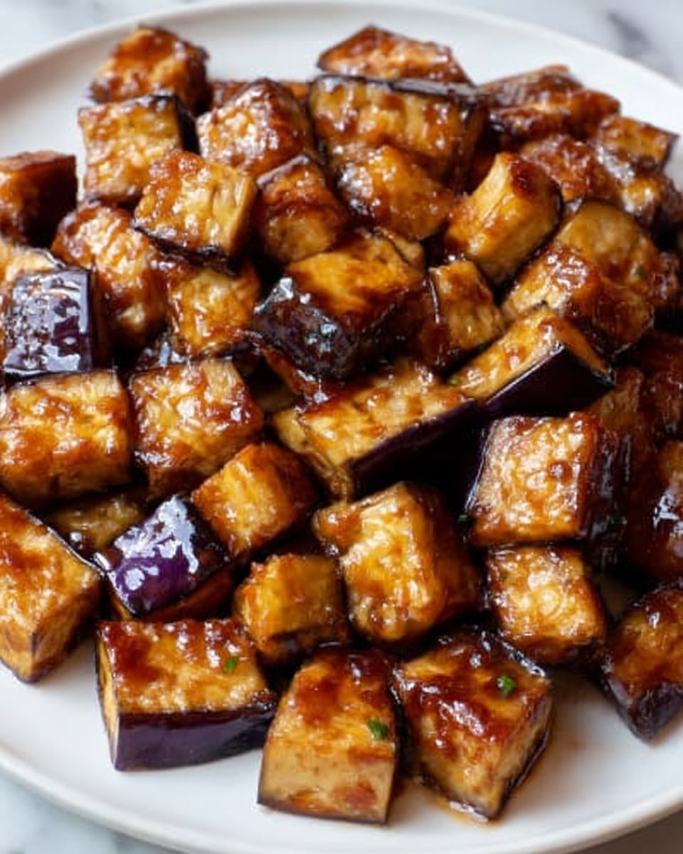 The image shows a white plate filled with many chunks of cooked eggplant. Each piece is golden brown with a shiny glaze of sauce that looks sticky and thick. The eggplant pieces are cut into medium-sized cubes with slightly charred edges that add a crispy texture. The color of the eggplant ranges from deep purple on the skin to a warm golden brown on the cooked parts, creating a rich contrast. The pieces are piled up closely together, covering the whole plate. The surface under the plate is a white marbled texture. photo taken with an iphone --ar 4:5 --v 7