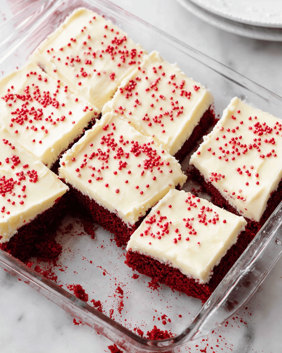 A square glass baking dish holds six cut pieces of red velvet cake with two layers: a dark red, moist cake base and a thick, smooth, creamy white frosting layer on top. The frosting covers each piece evenly and is decorated with small, round red sprinkles scattered across the surface. The dish sits on a white marbled surface, with some crumbs and smudges of red cake visible around the edges. In the background, part of a white plate is visible. photo taken with an iphone --ar 4:5 --v 7