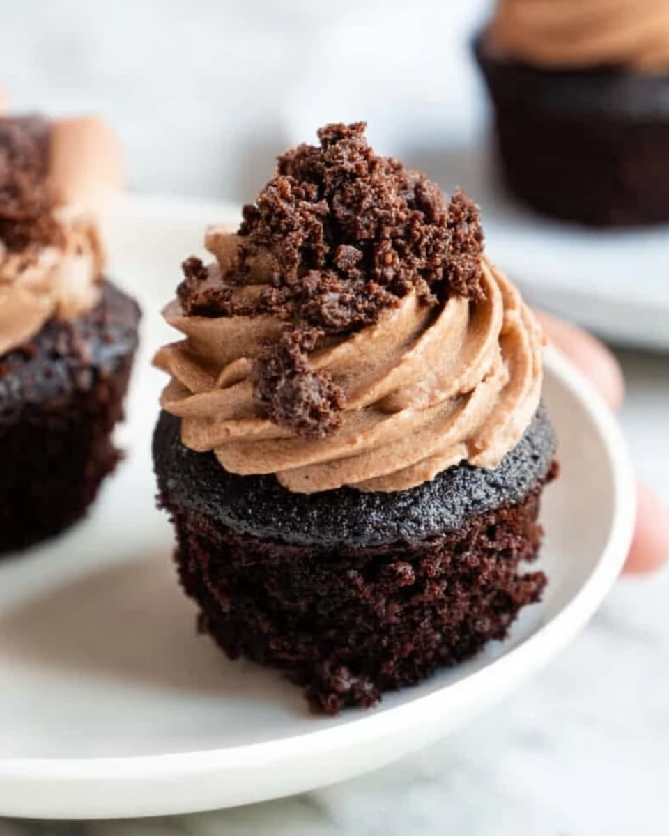 The image shows a close-up of a single dark chocolate cupcake placed on a white plate. The cupcake has two layers: the bottom layer is the dark, moist cake, and the top layer is a swirl of smooth, light brown chocolate frosting piped thickly. On top of the frosting, there are small, crumbly chocolate pieces scattered as decoration. The plate rests on a white marbled surface, and a woman's hand is slightly touching the edge of the plate in the background. Photo taken with an iphone --ar 4:5 --v 7