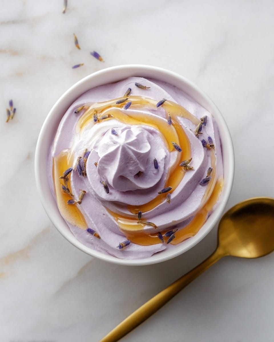 A white bowl filled with one smooth, thick layer of light purple whipped cream swirled into a peak in the center. On top of the whipped cream, there is a drizzle of golden honey flowing in a spiral pattern and scattered small dried lavender buds adding tiny purple and brown spots. The bowl is placed on a white marbled texture surface, with a gold spoon resting diagonally next to it. Photo taken with an iphone --ar 4:5 --v 7