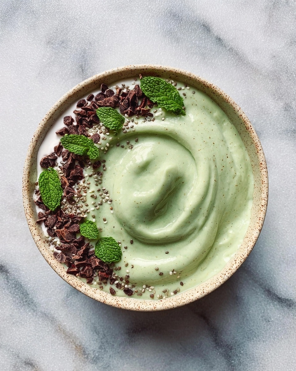 A bowl filled with a thick, smooth swirl of pale green smoothie sits centered on a white marbled surface. On the top left edge of the bowl, there is a thin layer of creamy white yogurt with a few small fresh mint leaves scattered on it. Around the yogurt are small clusters of dark brown cacao nibs and lightly sprinkled white chia seeds, which also spread faintly over the green smoothie. The bowl itself has a soft speckled beige rim that contrasts gently with the colorful contents inside. Photo taken with an iphone --ar 4:5 --v 7