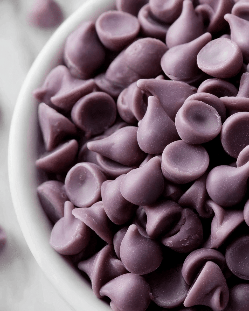 A close-up view of many small, purple, teardrop-shaped chocolate chips with smooth, shiny surfaces piled inside a white bowl, filling it almost to the top, with each chip showing a slight swirl at the pointed tip. The background shows a white marbled texture, slightly blurred to keep focus on the chips. Photo taken with an iphone --ar 4:5 --v 7