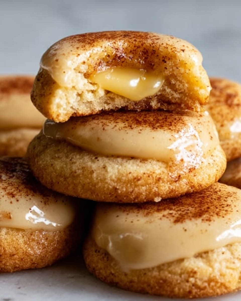 A close-up image of several small round cookies on a white marbled surface. Each cookie has a golden-brown outer crust with a slightly cracked texture, dusted lightly with a fine cinnamon powder on top. One cookie in the center is bitten, showing a smooth, creamy, pale yellow filling inside, contrasting with the crispy exterior. The cookies are arranged closely, overlapping each other slightly, creating a warm and inviting look. The photo taken with an iphone --ar 4:5 --v 7