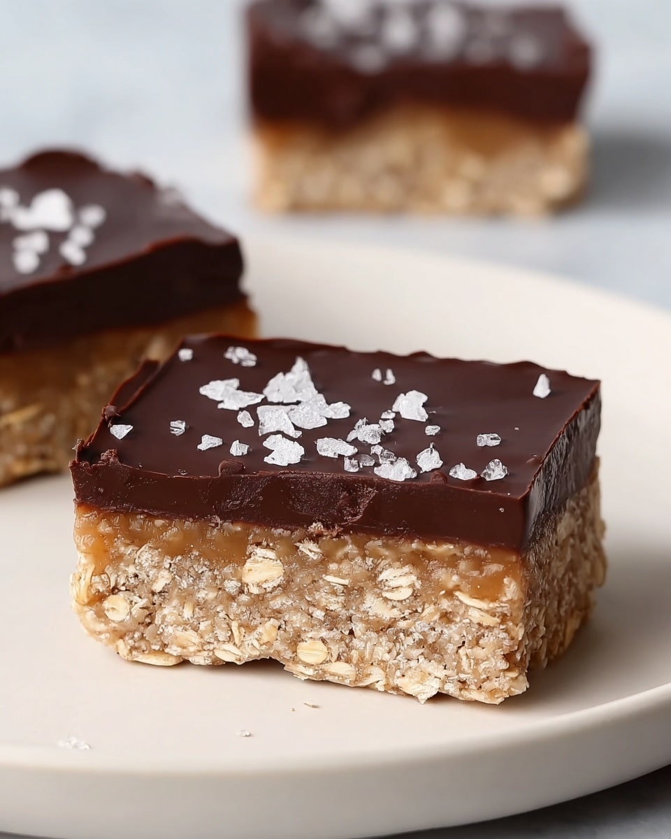 The image shows a close-up of a two-layered dessert bar placed on a white plate on a white marbled surface. The bottom layer is thick and textured with visible rolled oats and a light beige color, looking slightly crumbly but firm. The top layer is a smooth, glossy dark chocolate coating with a few coarse salt crystals sprinkled on it, adding a bit of shine and contrast. The bar is cut in a clean rectangular shape with the layers clearly defined and even. In the background, out of focus, is a similar dessert piece. Photo taken with an iphone --ar 4:5 --v 7