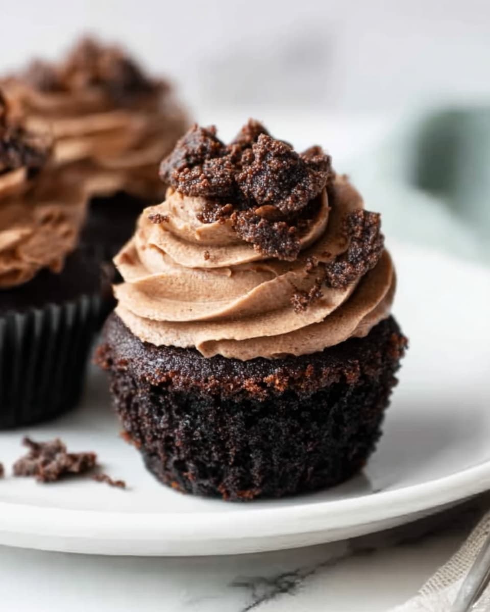 The image shows a close-up of a dark chocolate cupcake with two main layers: the base is a dense, moist, almost black cake with a rough texture, and the top is a thick swirl of smooth, rich milk chocolate frosting. The frosting is decorated with small, crunchy chocolate pieces scattered on top and around the base of the cupcake. The cupcake sits on a white plate that contrasts with its dark color, all placed on a white marbled surface. photo taken with an iphone --ar 4:5 --v 7