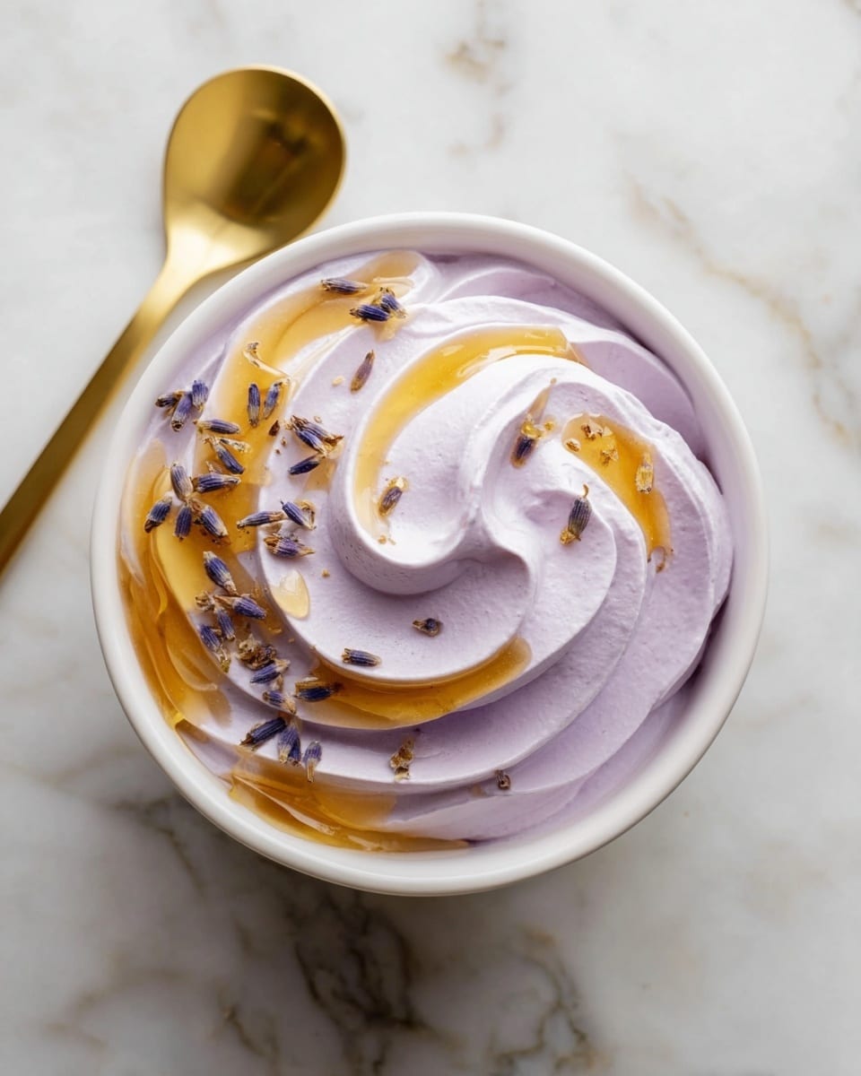 A white bowl filled with a single swirl of smooth, light purple whipped cream, topped with small dried lavender buds scattered evenly across the surface and a light drizzle of golden honey following the swirl's curve. The bowl rests on a white marbled texture surface, with a gold spoon placed diagonally to the right side of the bowl. The overall look is soft and elegant with gentle pastel and warm honey tones. photo taken with an iphone --ar 4:5 --v 7