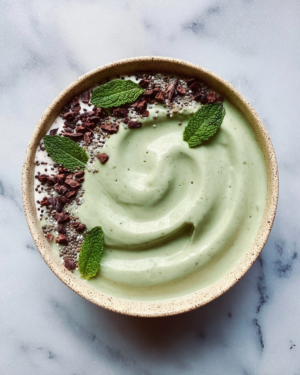 A white bowl filled with a smooth, thick swirl of green smoothie forms the main layer, slightly raised in the center. On the top left edge of the swirl, there is a small dollop of light creamy white yogurt or sauce spread thinly and unevenly. Scattered around the white layer on the upper left and top rim are dark brown cacao nibs, bright green fresh mint leaves, and tiny light beige sesame seeds sprinkled gently, creating contrast against the green and white. The bowl rests on a white marbled surface, adding subtle texture without drawing attention away from the bowl. Photo taken with an iphone --ar 4:5 --v 7