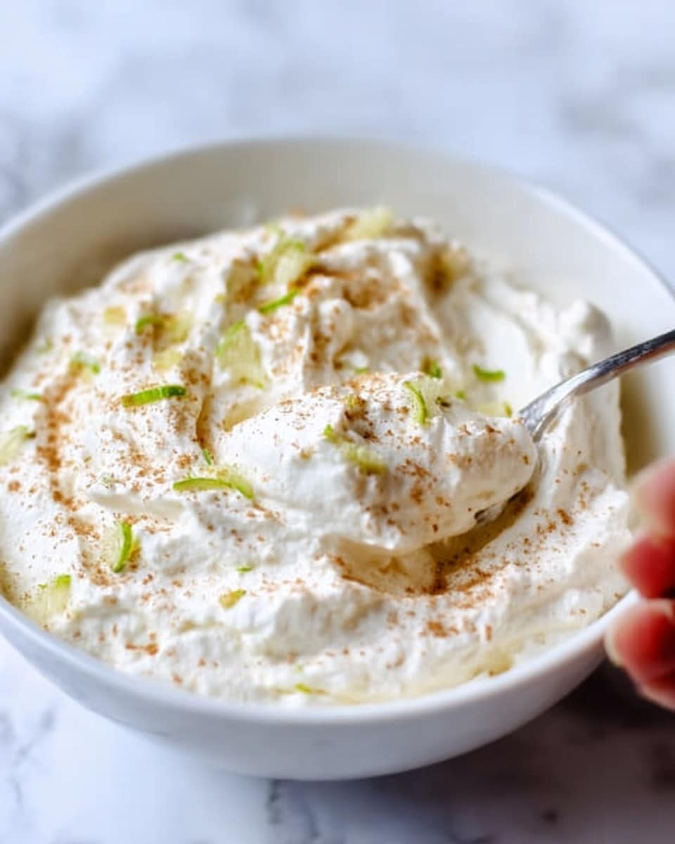 The image shows a white bowl filled with a fluffy, creamy white mixture that looks soft and airy. The top layer is sprinkled lightly with fine brown powder and small thin slivers of green zest, adding color contrast and texture to the smooth, whipped surface. A woman's hand is scooping some of the creamy mixture, showing its light, soft texture. The bowl is placed on a white marbled surface, and the lighting highlights the texture and fresh look of the food. photo taken with an iphone --ar 4:5 --v 7